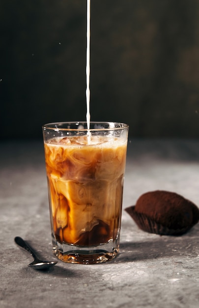 Kalter Kaffee in einem hohen Glas mit Milch- und Schokoladenkeksen an einer grauen dunklen Wand mit Kopienraum Milch wird in Kaffee gegossen