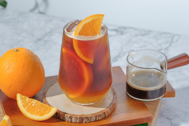 Kalter schwarzer Eiskaffee gemischt mit Orangensaft und Orangenscheiben im Glas auf Holzplatte auf weißem Hintergrund mit Naturblättern Silhouette Schatten zwei Töne Schicht frisches Sommergetränk am Morgen