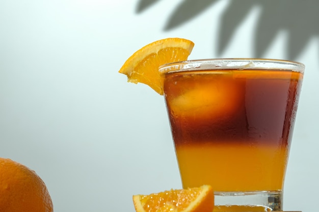 Kalter schwarzer Eiskaffee gemischt mit Orangensaft und Orangenscheiben im Glas auf Holzplatte auf weißem Hintergrund mit Naturblättern Silhouette Schatten zwei Töne Schicht frisches Sommergetränk am Morgen