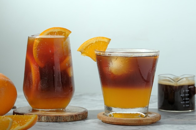 Kalter schwarzer Eiskaffee gemischt mit Orangensaft und Orangenscheiben im Glas auf Holzplatte auf weißem Hintergrund mit Naturblättern Silhouette Schatten zwei Töne Schicht frisches Sommergetränk am Morgen