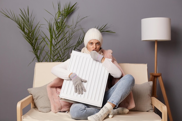 Kaltgefrorene Frau in Mantel und Hut sitzt im kalten Wohnzimmer und leidet unter mangelnder Hitze, die auf ihre Hände weht, um sich warm zu machen, während sie auf dem Sofa sitzt