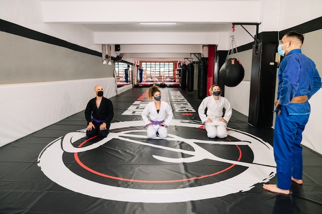 Kampfsportstudenten mit Kimonos auf der Bodenmatte im Fitnessstudio hören den Anweisungen ihres Lehrers zu und tragen wegen der Covid 19 Gesichtsmasken