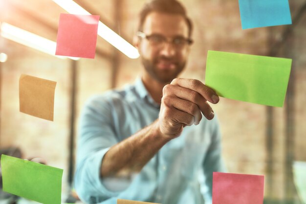 Kanban board jovem e bonito homem barbudo em óculos usando adesivos coloridos e sorrindo enquanto