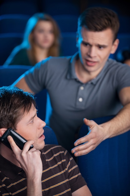 Kannst du aufhören zu reden? Junger Mann bittet darum, den anderen Mann zu stoppen, der am Handy telefoniert, während er im Kino einen Film sieht