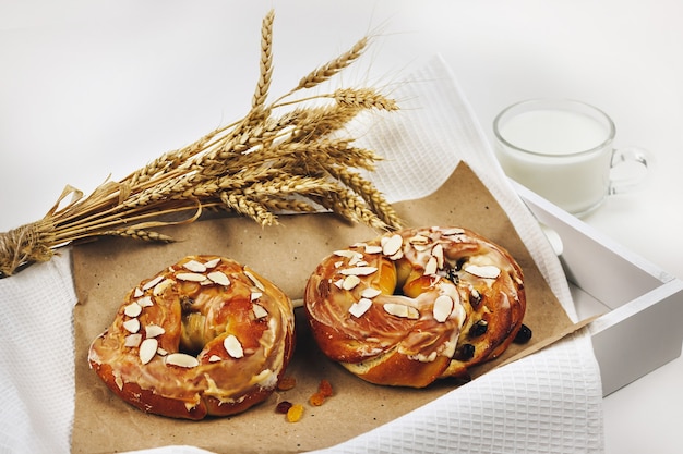 Foto karamellbrötchen mit rosinen und mandeln liegen auf kraftpapier in einem tablett. daneben ein strauß weizen- und roggenohren, ein becher mit milch auf konkretem hintergrund, selektiver fokus.