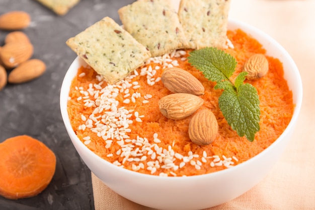 Karottencremesuppe mit Sesam und Snacks in der weißen Schüssel auf einem schwarzen Betonhintergrund. Seitenansicht, Nahaufnahme.