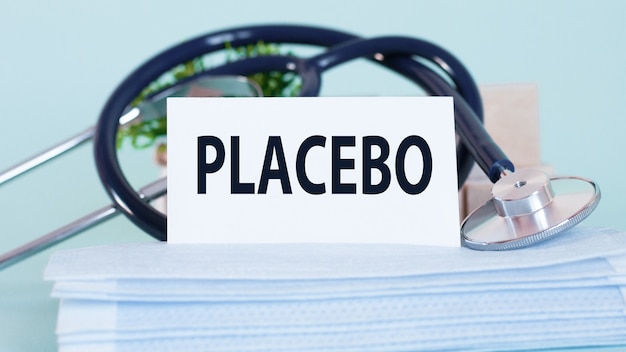 Karte mit Wort Placebo, Stethoskop, Gesichtsmasken und Blume auf Tisch. Persönliche Schutzausrüstung auf weichem blauem Hintergrund. Medizin- und Gesundheitskonzept.