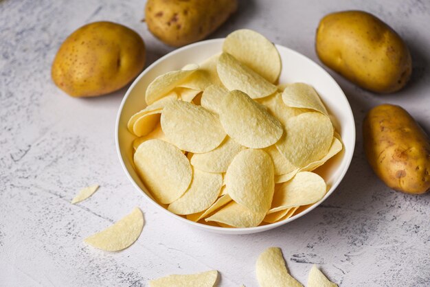 Kartoffelchips-Snack auf weißer Schüssel Knusprige Kartoffelchips auf dem Küchentisch und frische rohe Kartoffeln