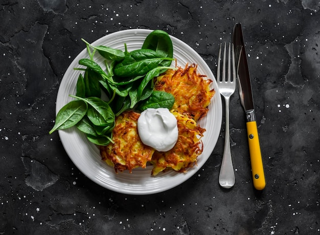 Kartoffellatkes mit saurer Sahne und Spinatsalat auf einer Draufsicht des dunklen Hintergrundes