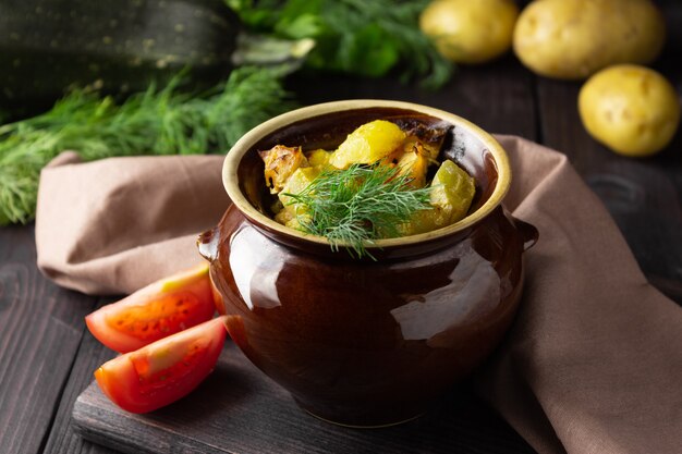 Foto kartoffeln, hühnchen und zucchini in einem tontopf auf dunklem hintergrund gebacken.