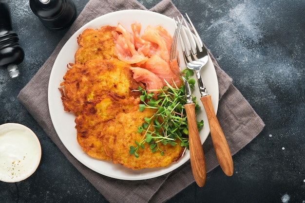 Kartoffelpfannkuchen Gebratene hausgemachte Kartoffelpfannkuchen oder Latkes mit cremefarbenen Frühlingszwiebeln, Mikrogrün, roter Lachs und Soße in rustikalem Teller auf altem schwarzem Tischhintergrund Rustikale Ansicht von oben