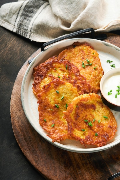 Kartoffelpfannkuchen Gebratene hausgemachte Kartoffelpfannkuchen oder Latkes mit Sahne und Frühlingszwiebeln in rustikaler Platte auf altem schwarzem Holztischhintergrund Rustikaler Stil Gesundes Essen Draufsicht