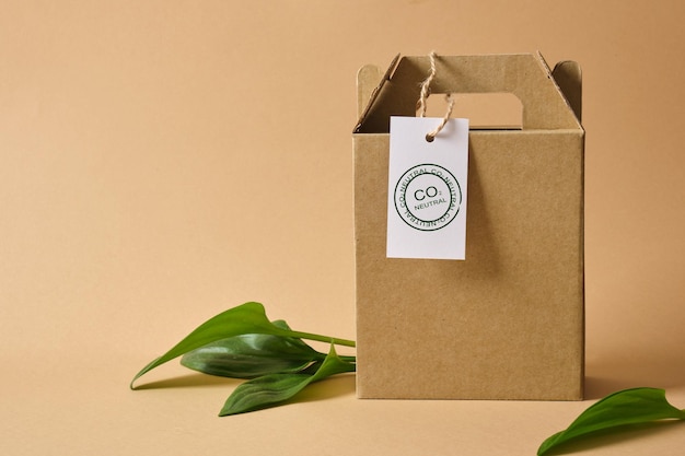 Foto karton mit griffen für lieferung oder geschenk, blätter und weißes etikett mit co2-neutralon auf braunem hintergrund umweltfreundliche verpackung, papierrecycling, zero waste, naturproduktkonzept. platz kopieren.