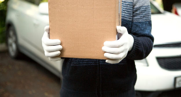 Foto karton mit platz für text in den händen eines männlichen kuriers in weißen handschuhen. kurier auf dem hintergrund eines weißen autos.