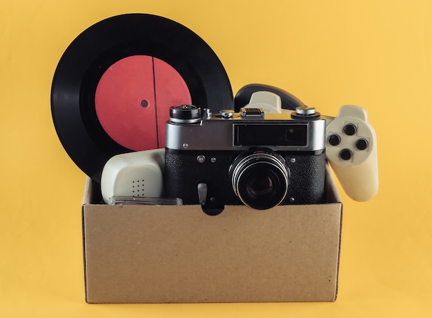 Karton mit Retro-Filmkamera, Gamepad, Schallplatte, Telefonröhre auf Gelb