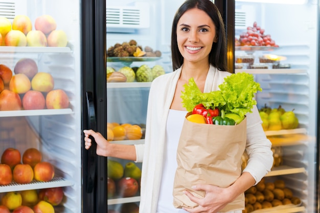 Kauf der frischesten Produkte. Schöne junge Frau, die Einkaufstasche mit Früchten hält und lächelt, während sie im Lebensmittelgeschäft in der Nähe des Kühlschranks steht