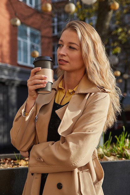 Kaukasische attraktive Frau im beigen Trenchcoat, der draußen Tasse Kaffee hält