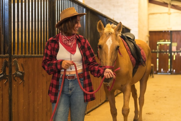 Kaukasische Cowgirl-Frau, die Spaß hat und mit einem Pferd in einem Stall lächelt, amerikanischer Cowboyhut, rotes kariertes Hemd und Jeans