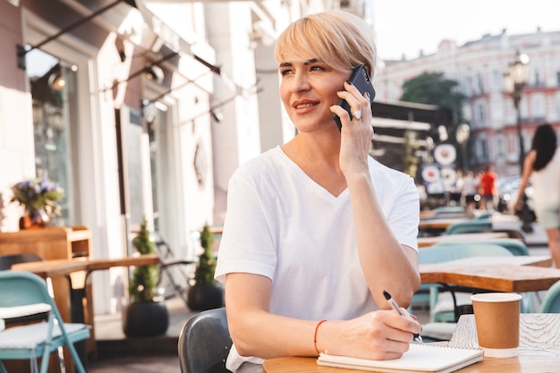 Kaukasische erfreute Frau, die weißes T-Shirt trägt, das im Straßencafé sitzt, während in Notizbuch schreibt und auf Handy spricht