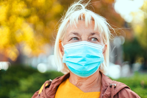Kaukasische erwachsene blonde Frau 50 Jahre alt in hellem Gewand und schützender medizinischer Atemschutzmaske, die sich auf dem Parkhintergrund umschaut