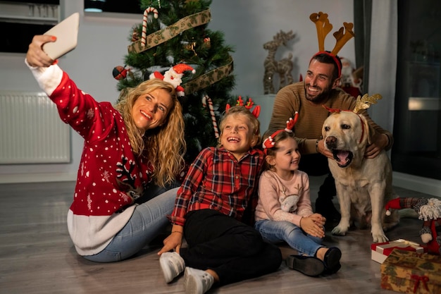 Kaukasische Familie mit Weihnachtsstirnbändern und einem Labrador-Hund, der ein Selfie mit dem Smartphone in der Nähe des Weihnachtsbaums macht