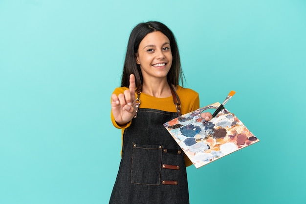 Kaukasische Frau des jungen Künstlers, die eine Palette lokalisiert auf blauem Hintergrund hält, die einen Finger zeigt und anhebt