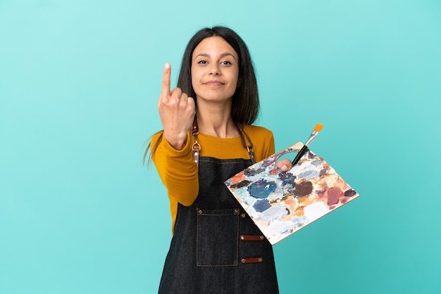 Kaukasische Frau des jungen Künstlers, die eine Palette lokalisiert auf blauem Hintergrund hält, die kommende Geste tut