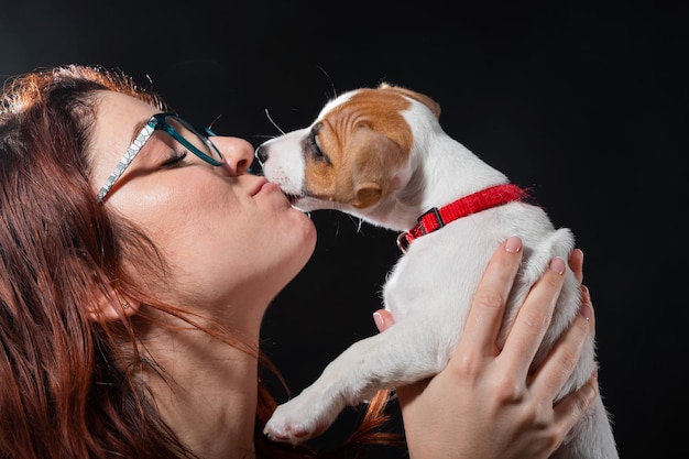 Kaukasische Frau, die einen Jack-Russell-Terrier-Welpen auf schwarzem Hintergrund küsst
