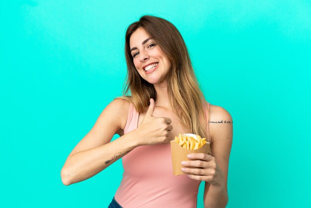 Kaukasische Frau, die gebratene Chips auf blauem Hintergrund hält und eine Geste mit dem Daumen nach oben gibt