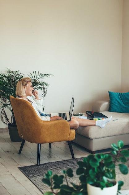 Kaukasische Frau, die im Sessel sitzt und am Telefon spricht, während Hausaufgaben auf dem Laptop machen