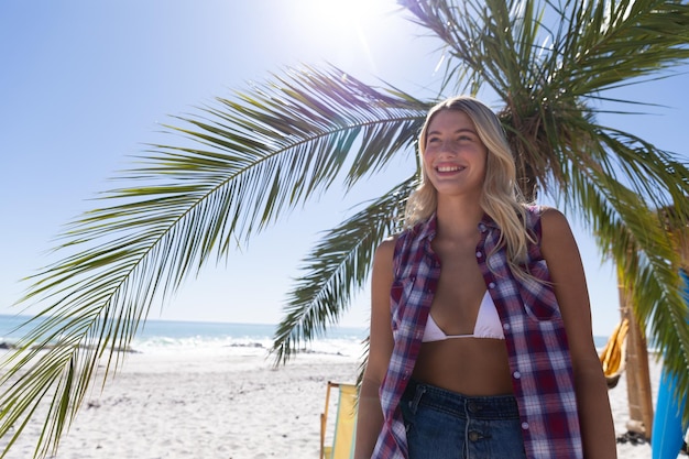 Kaukasische Frau genießt die Zeit am Strand an einem sonnigen Tag, steht neben einer Palme und lächelt mit dem Meer im Hintergrund