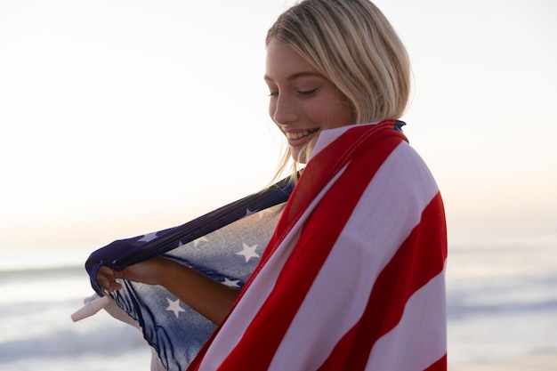 Kaukasische Frau genießt die Zeit am Strand bei Sonnenuntergang und bedeckt sich mit einer US-Flagge mit blauem Meer im Hintergrund