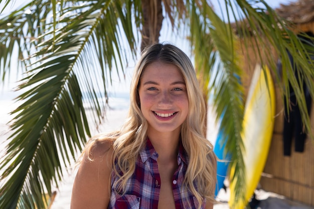 Kaukasische Frau genießt die Zeit am Strand, steht neben einer Palme, blickt in die Kamera und lächelt