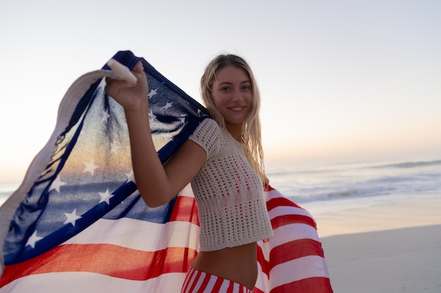 Kaukasische Frau genießt die Zeit am Strand während eines Sonnenuntergangs und hält und schwenkt eine US-Flagge mit blauem Himmel im Hintergrund