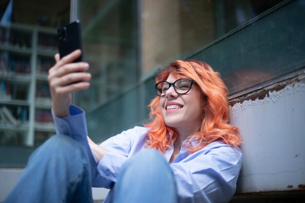 Kaukasische Frau mit Brille, die mit einem Lächeln im Freien auf ihren Handybildschirm schaut