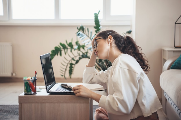 Kaukasische Frau mit den lockigen Haaren, die es satt hat, Brillen und Kopfhörer zu tragen, die Hausaufgaben am Laptop machen