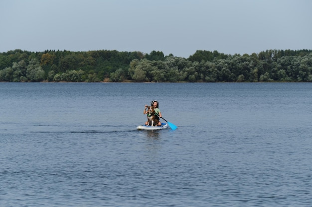 Kaukasische Frau mit Dreadlocks sitzt an Bord im See und rudert mit Ruder Australian Shepherd