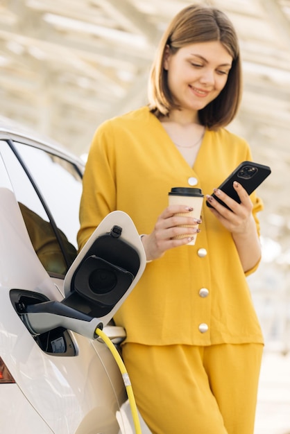 Kaukasische Frau mit Handy und Kaffeetasse, die in der Nähe des aufladenden Elektroautos steht