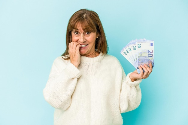 Foto kaukasische frau mittleren alters, die banknoten einzeln auf blauem hintergrund hält und fingernägel beißt, nervös und sehr ängstlich.