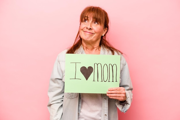 Kaukasische Frau mittleren Alters, die das Plakat "Ich liebe Mama" isoliert auf rosafarbenem Hintergrund hält und davon träumt, Ziele und Zwecke zu erreichen