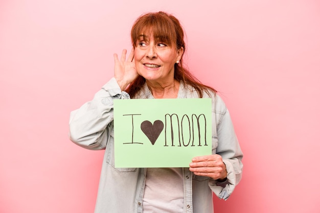 Kaukasische Frau mittleren Alters, die das Plakat "Ich liebe Mama" isoliert auf rosafarbenem Hintergrund hält und versucht, einen Klatsch zu hören