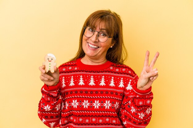 Kaukasische Frau mittleren Alters, die ein Weihnachtsplätzchen einzeln auf gelbem Hintergrund hält, freudig und sorglos, das ein Friedenssymbol mit den Fingern zeigt.