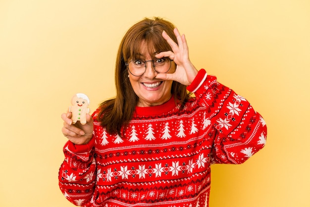 Kaukasische Frau mittleren Alters, die ein Weihnachtsplätzchen hält, das auf gelbem Hintergrund isoliert ist, aufgeregt, die Geste in Ordnung auf dem Auge zu halten.