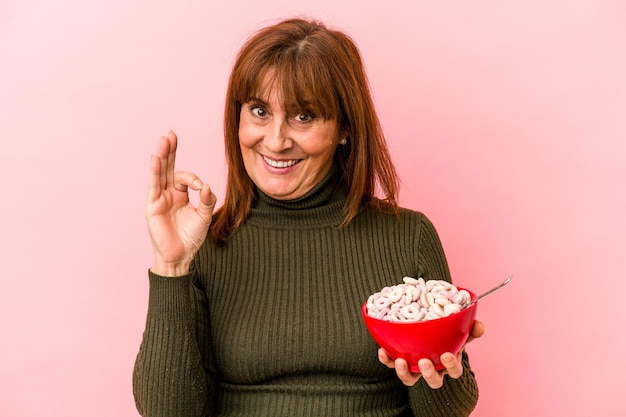 Kaukasische Frau mittleren Alters, die eine Schüssel mit Müsli einzeln auf rosafarbenem Hintergrund hält, fröhlich und selbstbewusst, die eine ok Geste zeigt.