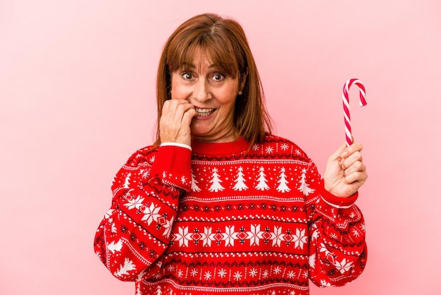 Kaukasische Frau mittleren Alters, die einen Weihnachtsstock einzeln auf rosafarbenem Hintergrund hält und Fingernägel beißt, nervös und sehr ängstlich.