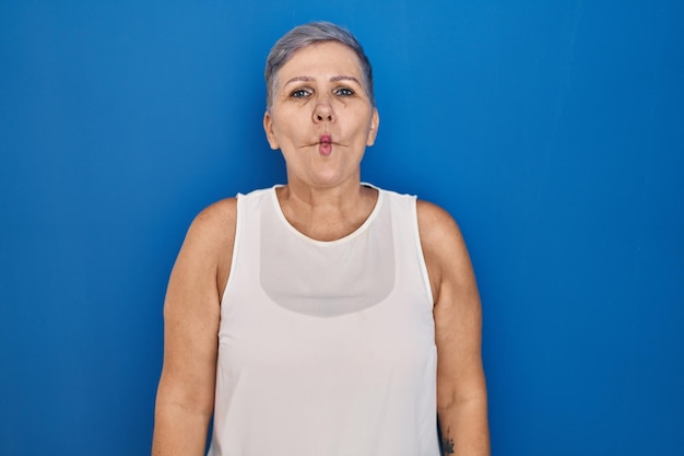 Kaukasische Frau mittleren Alters, die über blauem Hintergrund steht und Fischgesicht mit Lippen, verrückter und komischer Geste macht. lustiger Ausdruck.
