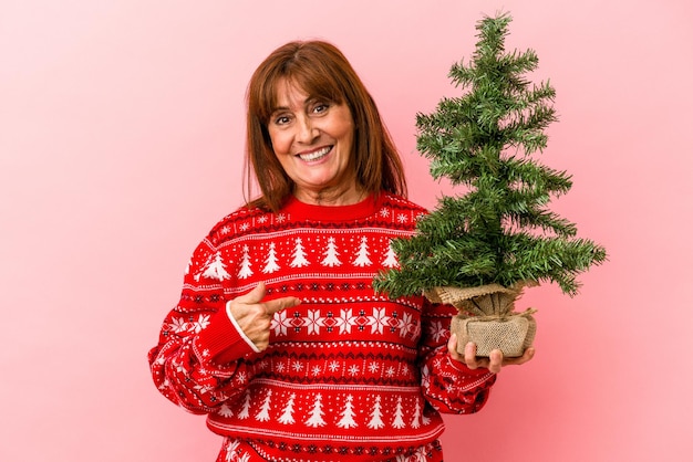 Kaukasische Frau mittleren Alters, die Weihnachtsbaum einzeln auf rosafarbenem Hintergrund hält, die mit der Hand auf einen Hemdkopierraum zeigt, stolz und selbstbewusst