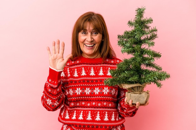 Kaukasische Frau mittleren Alters, die Weihnachtsbaum isoliert auf rosafarbenem Hintergrund hält, lächelt fröhlich und zeigt Nummer fünf mit den Fingern.