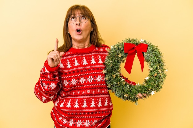 Kaukasische Frau mittleren Alters feiert Weihnachten mit Kranz isoliert auf gelbem Hintergrund, der mit geöffnetem Mund nach oben zeigt.