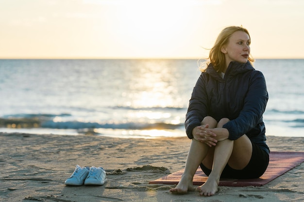 Kaukasische Frau sitzt auf einer Yogamatte mit entspanntem Ausdruck und ihren Schuhen neben ihr am Strand